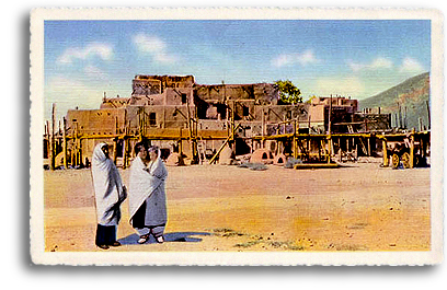 Two Native American (Indian) women stand talking, with the historic Taos Pueblo behind them. Their distance from the tiered dwellings offers a unique perspective on the majesty of the Pueblo and its innovative design and construction. This same pueblo-style is used today in much of the building of new homes and structures in the Taos area.